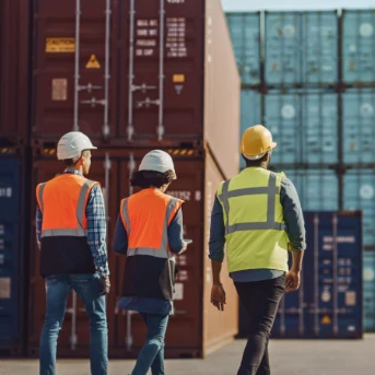 Two men & one woman in hard hats/safety vests walk away through a container terminal, with tall stacks of red/blue shipping containers behind.